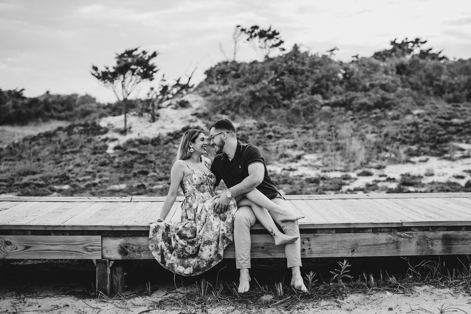 Black and white photo of a couple sitting close on a wooden boardwalk near sand dunes, sharing a romantic moment.