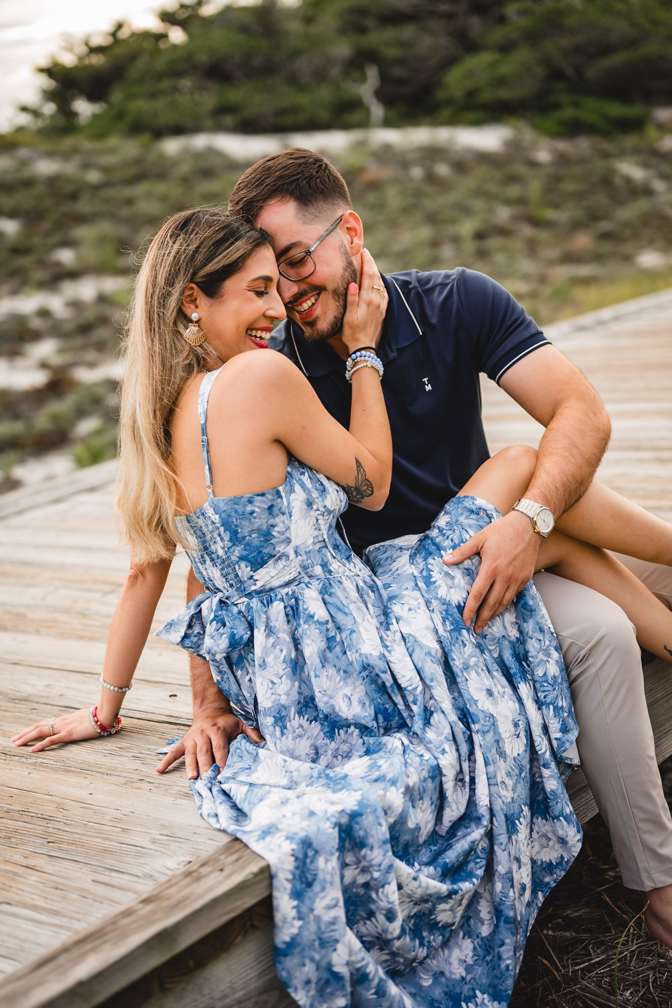Couple sitting on wooden boardwalk, woman in blue floral dress laughing as man leans in close near beach.