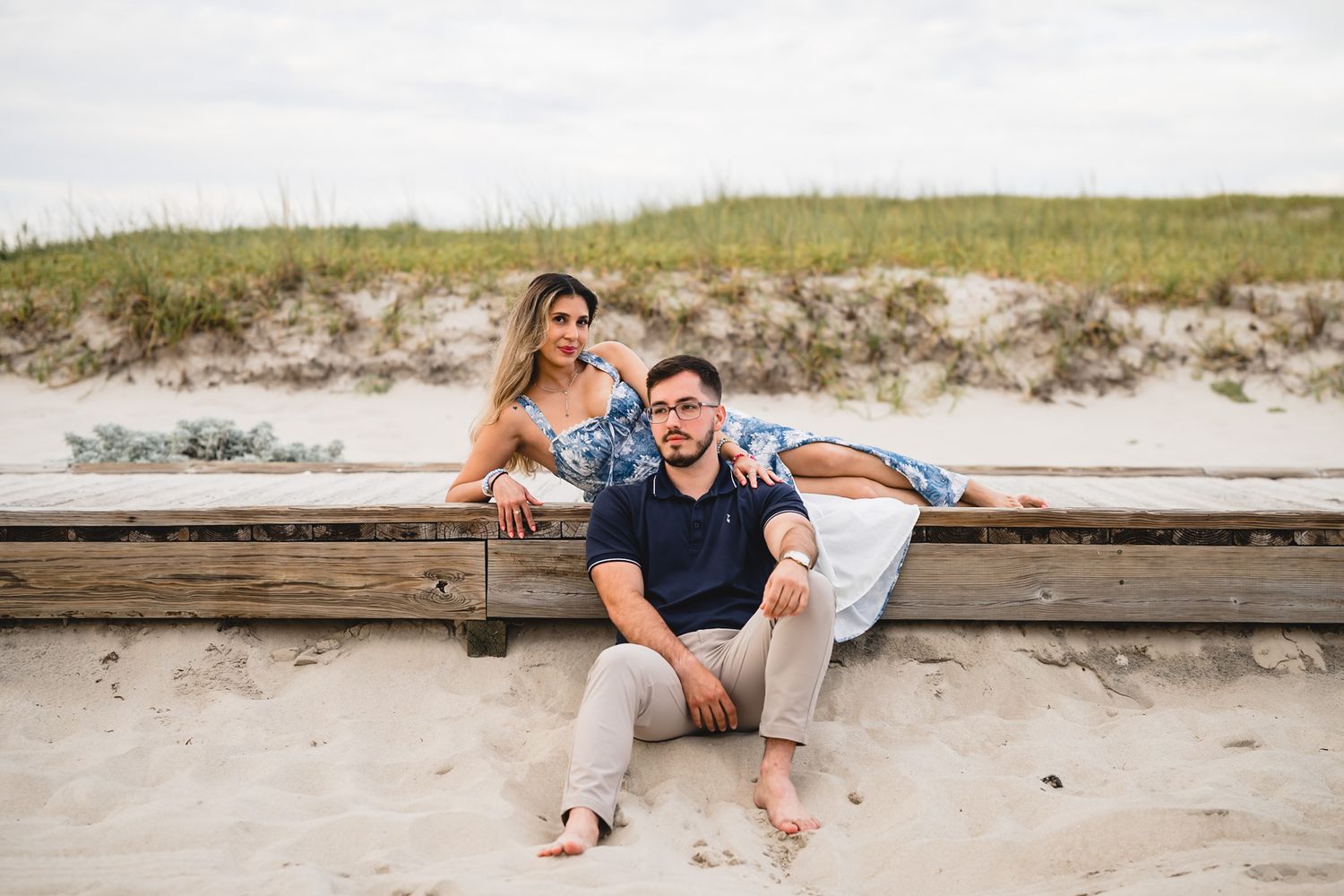 Couple posing on sandy beach boardwalk, woman leaning on bench, man sitting on sand, dunes in background.