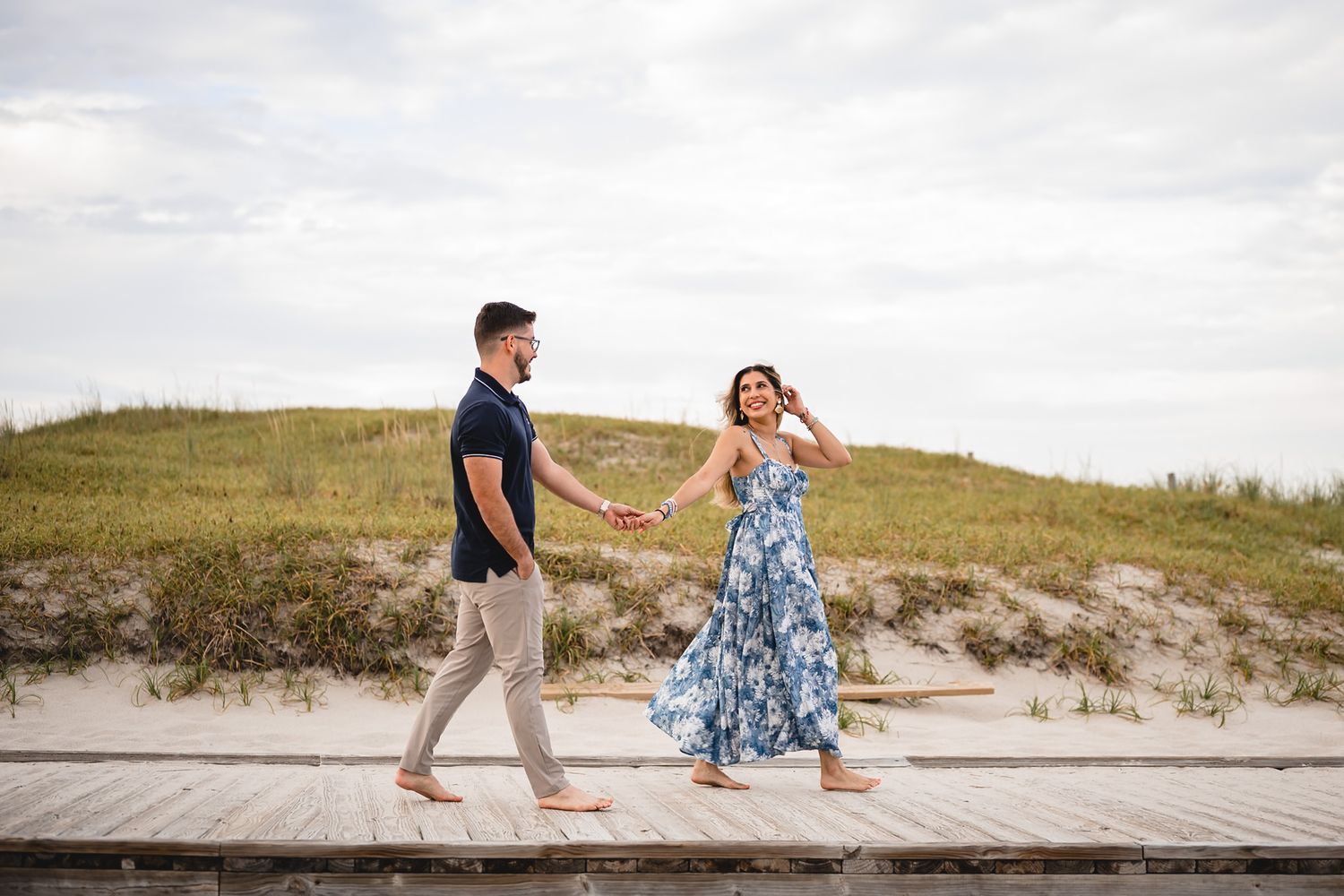 Happy couple holding hands walking on beach boardwalk, woman in blue floral maxi dress, sandy dunes behind.