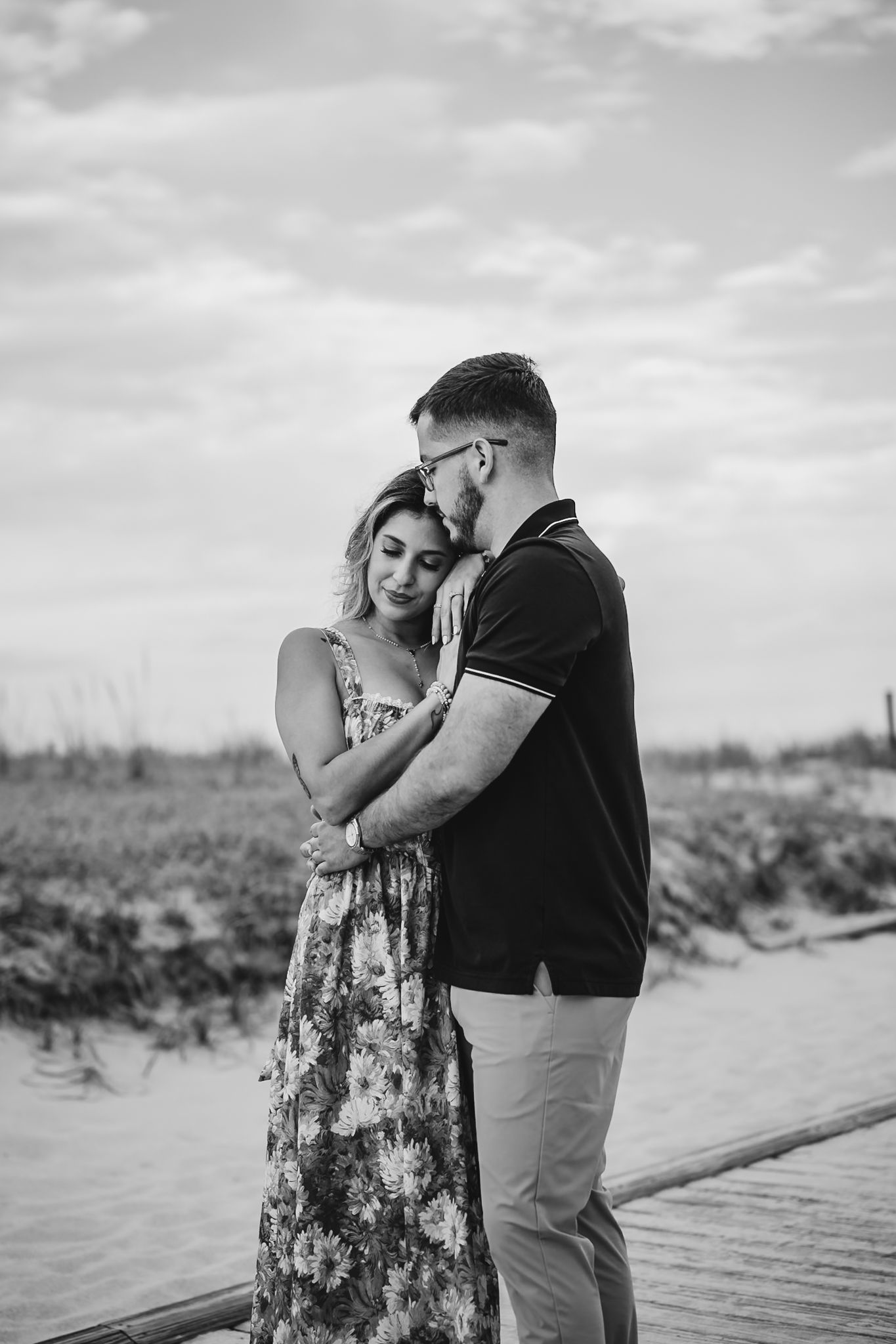 Black and white photo of a couple embracing tenderly on a wooden dock near a marsh under a cloudy sky.