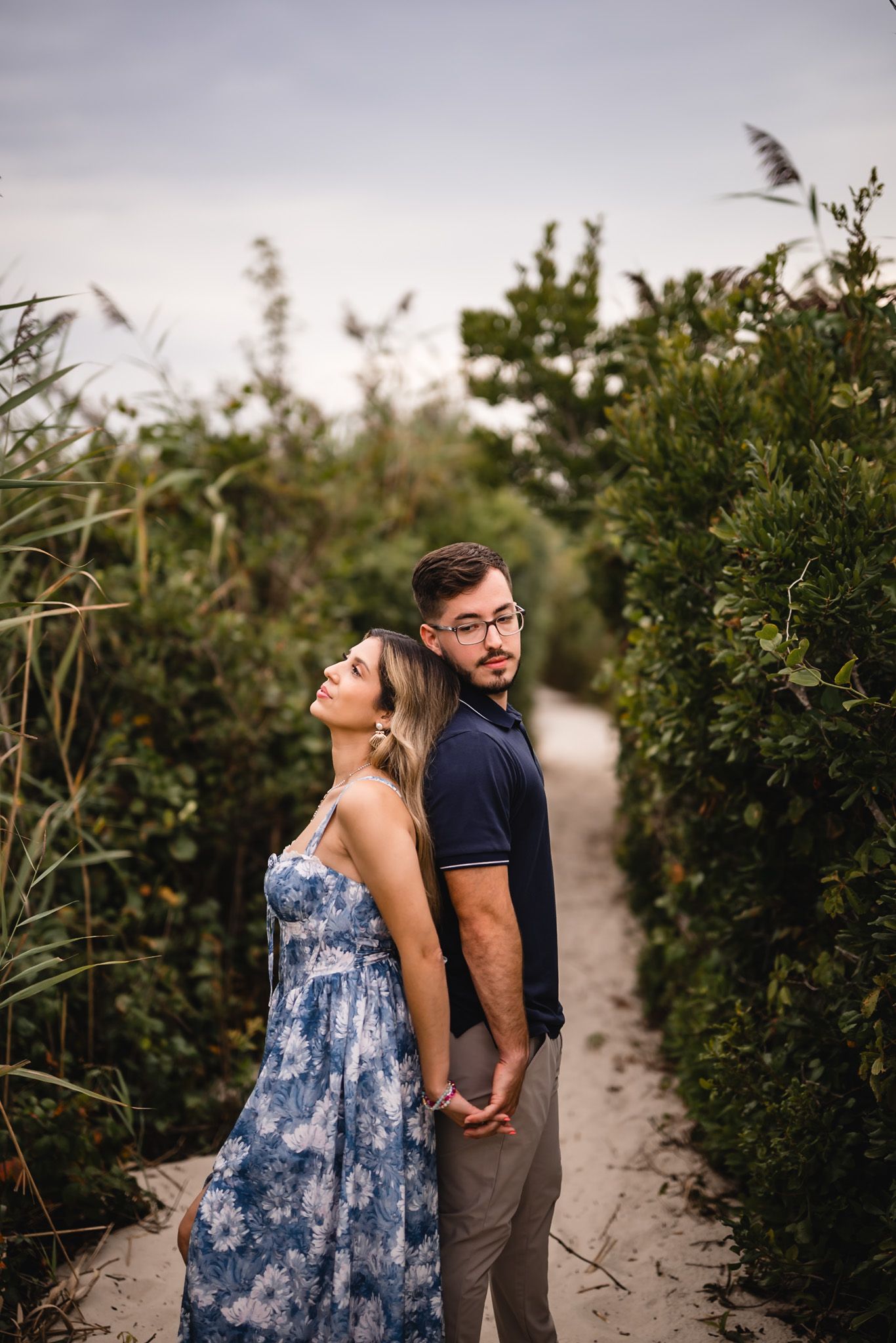Couple holding hands on a sandy path surrounded by lush green bushes, woman in floral dress leaning back on mans shoulder.
