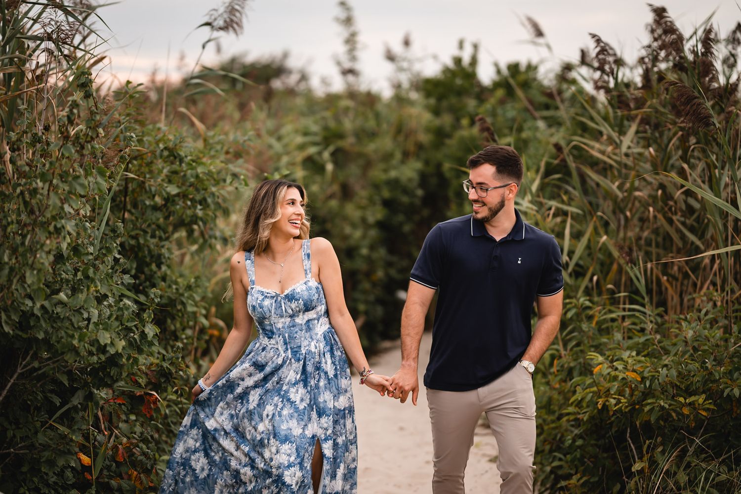 Couple holding hands walking on sandy path surrounded by lush greenery, woman in floral dress, man in navy polo.