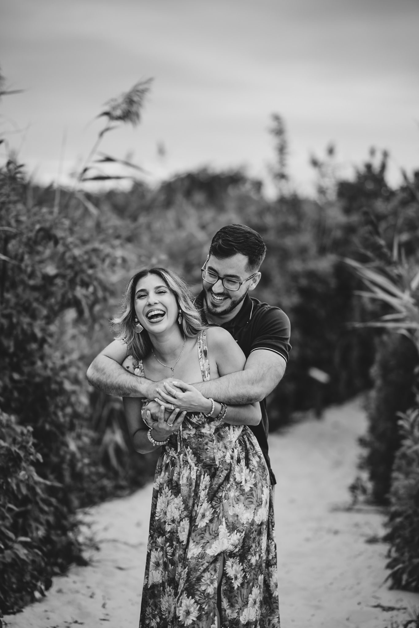Black and white photo of a laughing couple embracing on a sandy path surrounded by lush greenery.