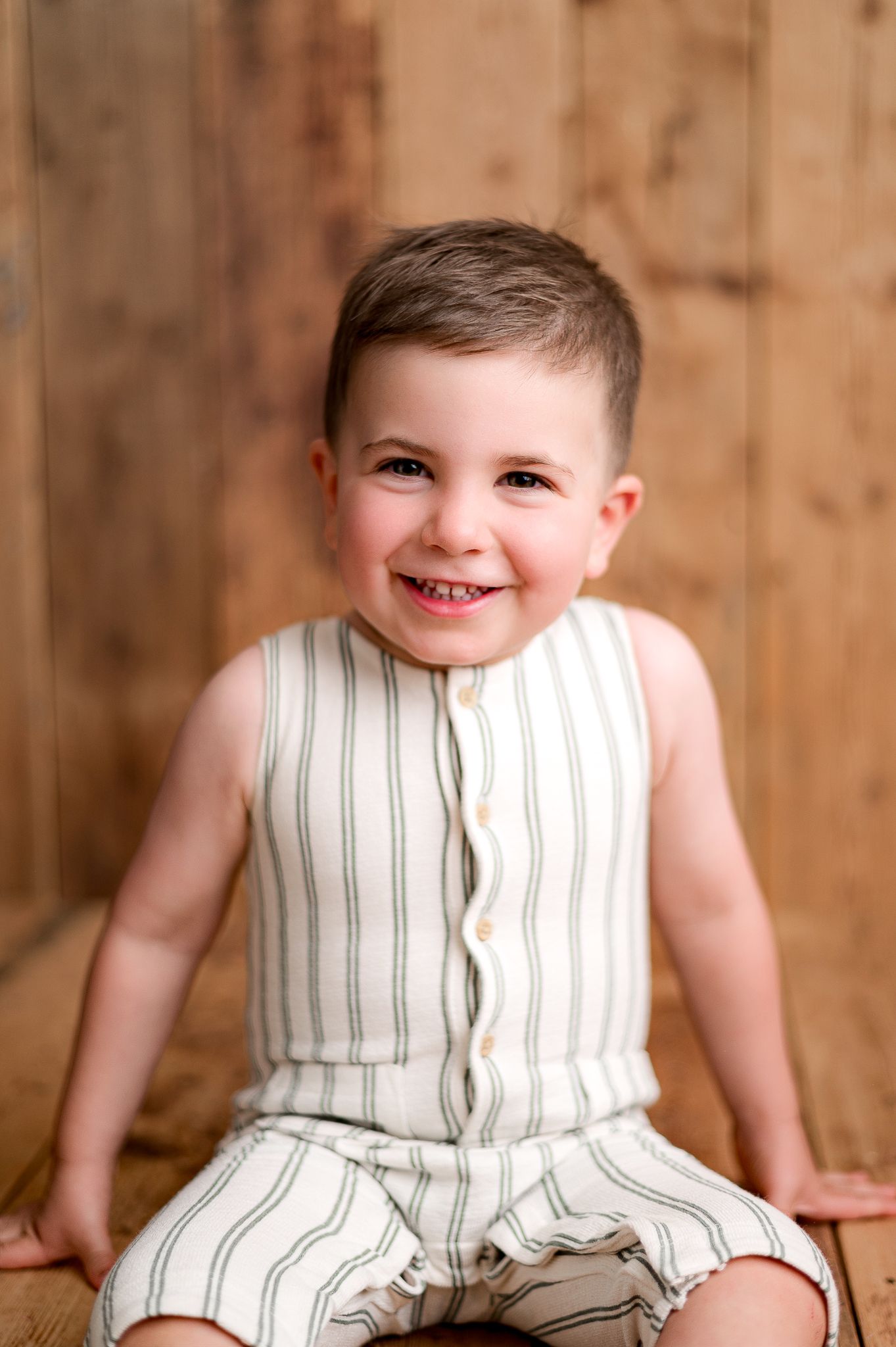 Smiling toddler boy in striped linen romper sitting on wooden floor against rustic wood backdrop.