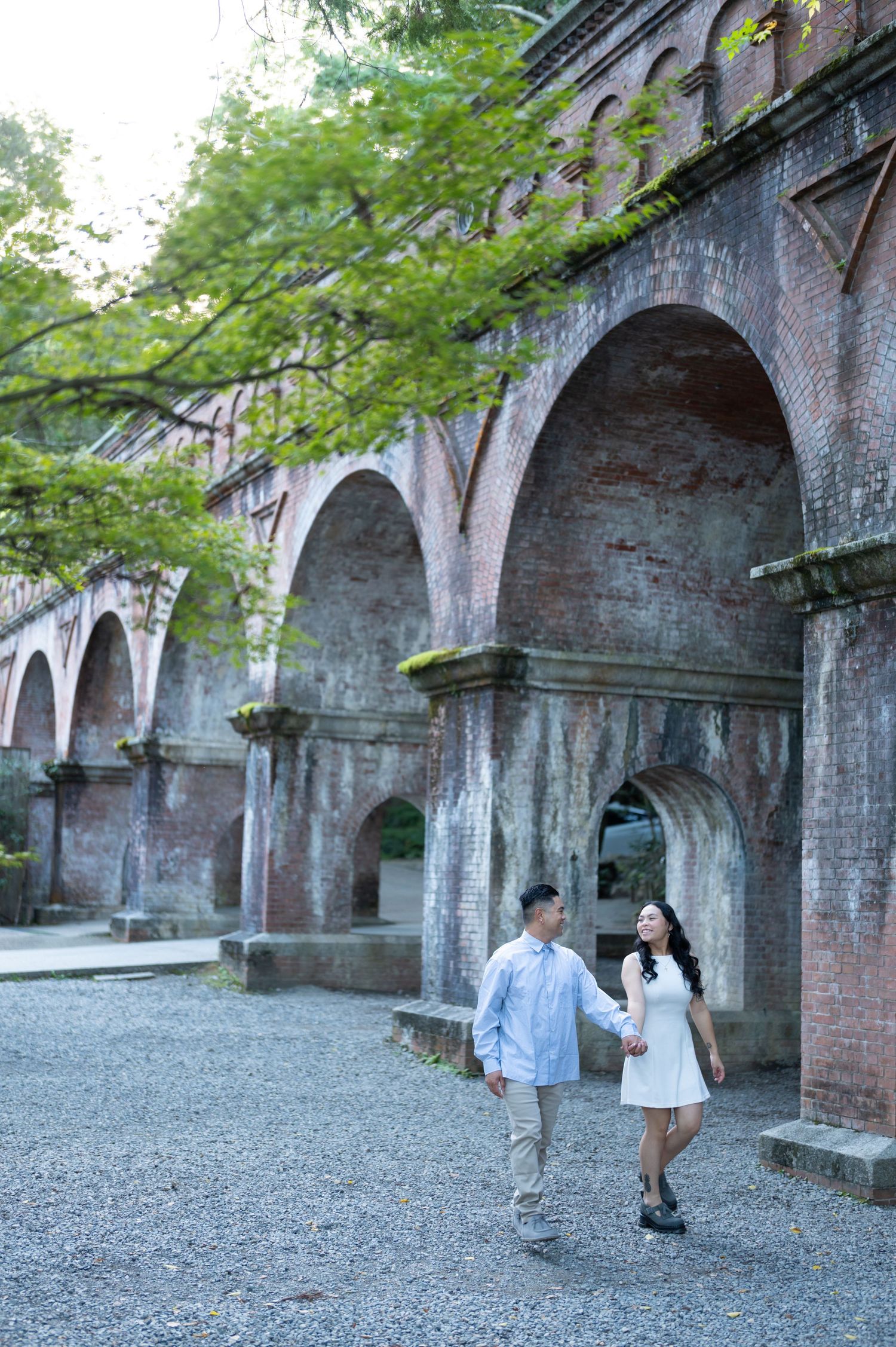 Couple holding hands walking beside historic red brick arched aqueduct surrounded by lush green trees.