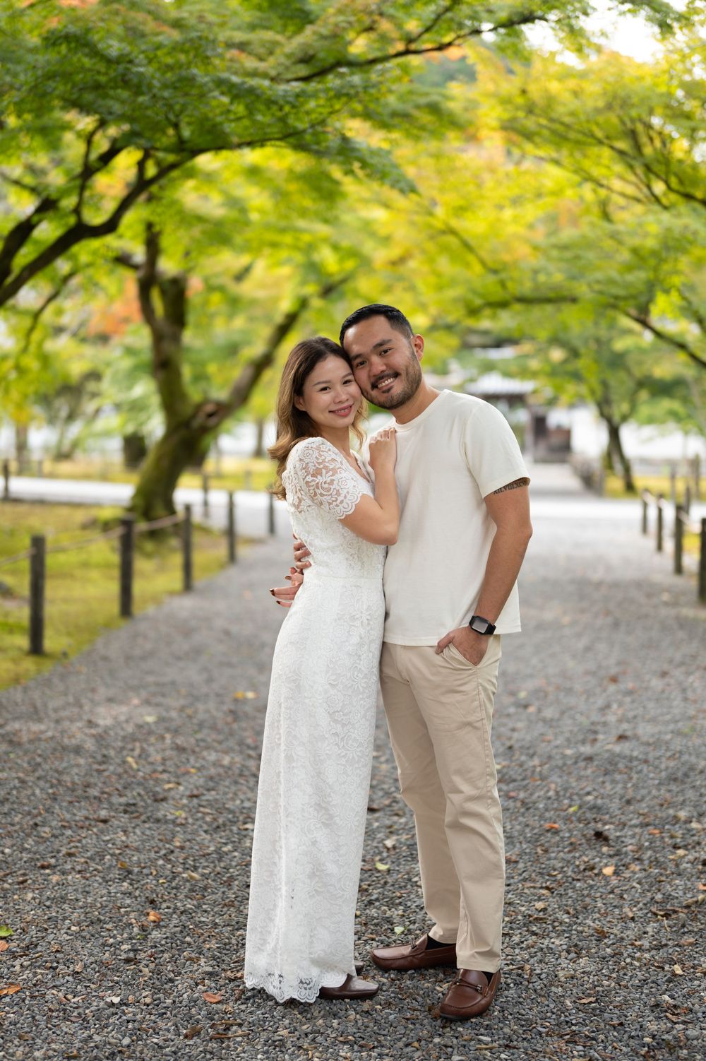 Couple posing together on a gravel path surrounded by lush green trees, woman in white lace dress, man in casual beige outfit.