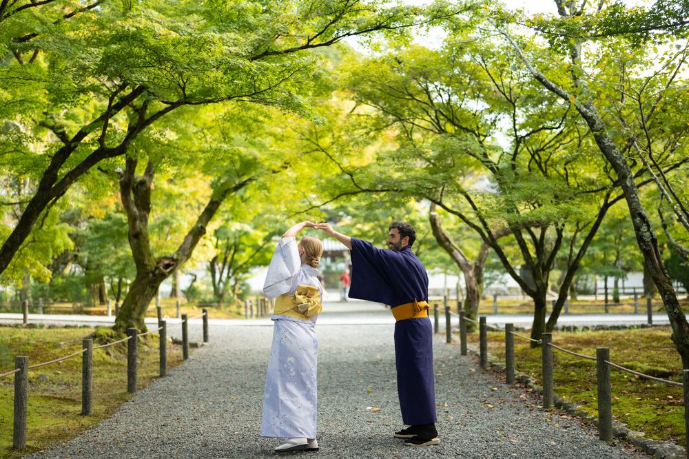 Two people in traditional Japanese kimono bow to each other on a tree-lined garden path in Japan.