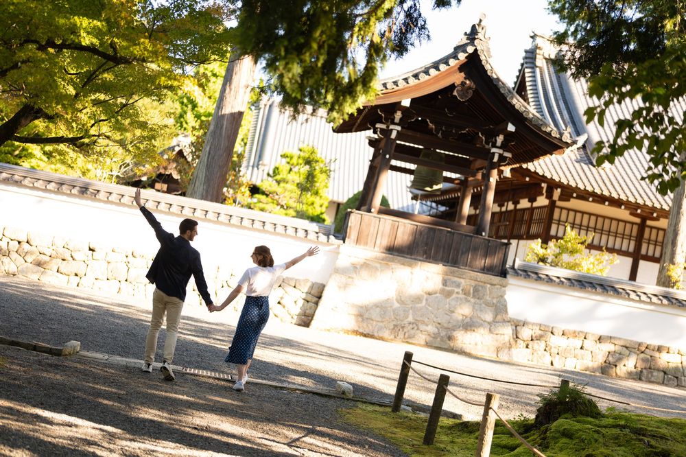 Couple holding hands walking toward a traditional Japanese wooden gate surrounded by lush green trees on a sunny day.