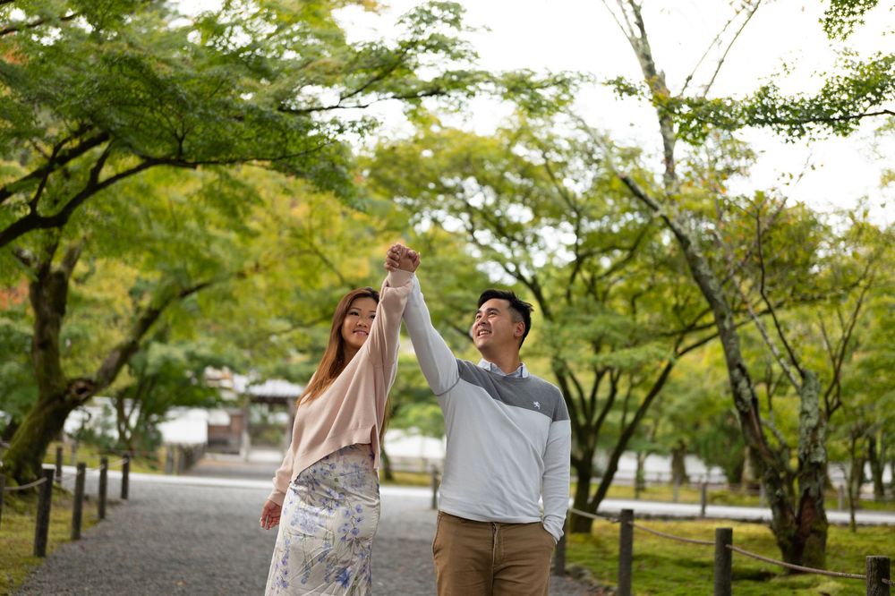 A joyful couple holds hands and dances together on a tree-lined park path in a lush green garden.