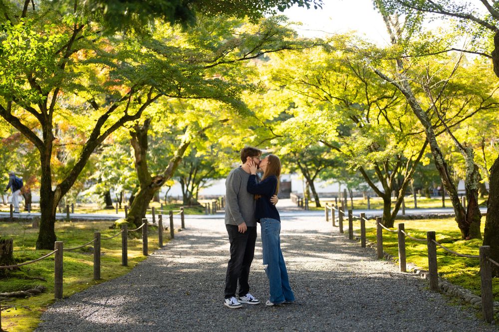 A couple shares a romantic kiss on a gravel path lined with lush green trees in a serene Japanese garden.