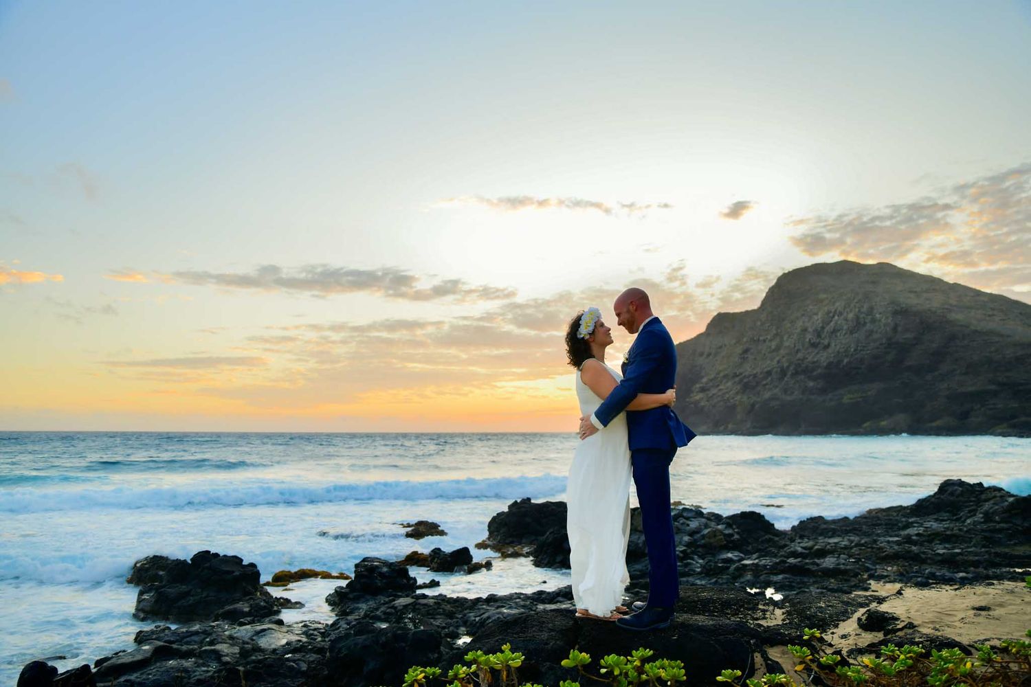 Couple shares romantic embrace at rocky ocean shore during golden sunset with mountain backdrop at their beach wedding.