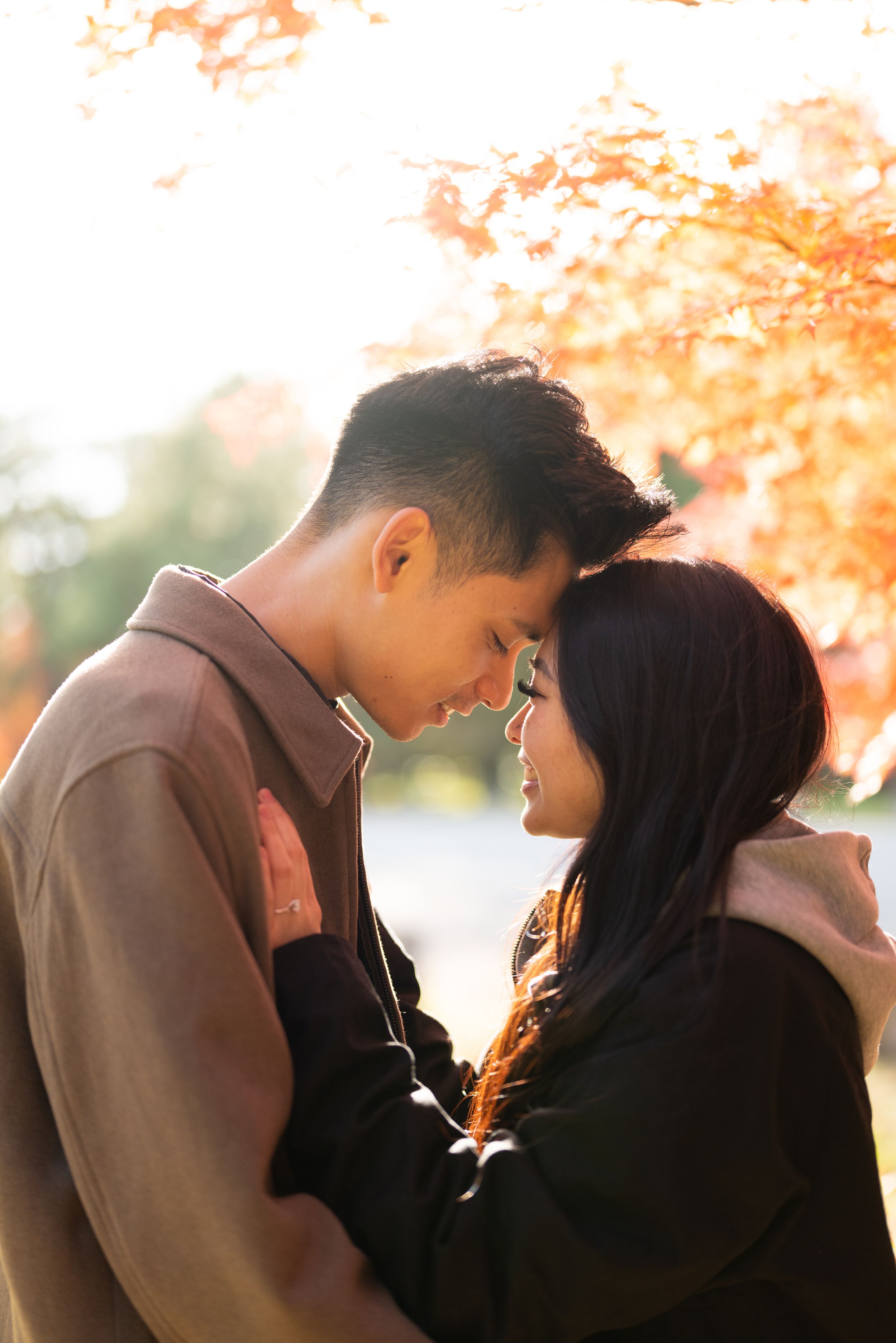 Couple embracing with foreheads touching under vibrant orange autumn foliage in soft, warm sunlight.