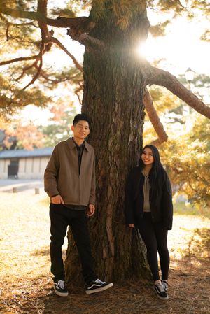 Smiling couple in casual jackets and sneakers standing on either side of a large pine tree in a sunlit autumn park.