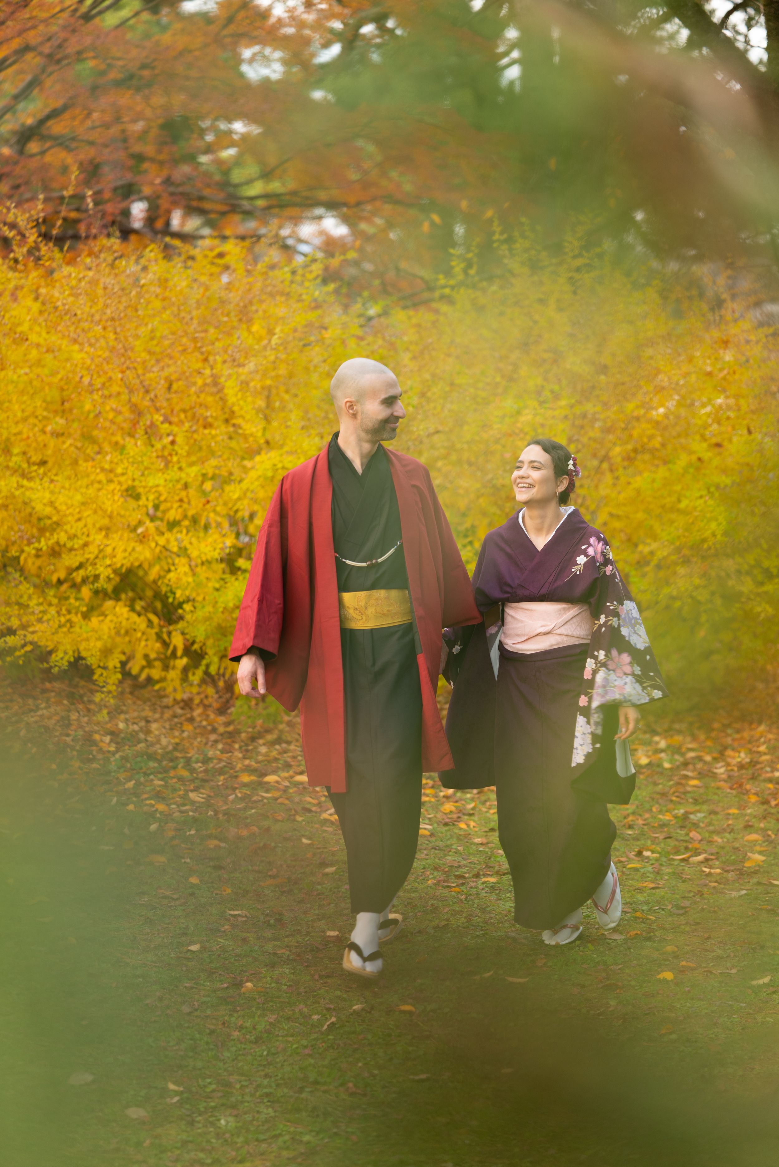Smiling couple in traditional Japanese kimono walking hand in hand through a park lined with golden yellow autumn foliage.