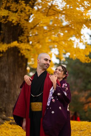 Smiling couple in traditional Japanese kimono embracing beneath a golden ginkgo tree surrounded by vibrant yellow autumn leaves.