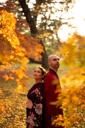 Couple in traditional Japanese kimono standing together beneath glowing orange autumn maple trees in a sunlit park.