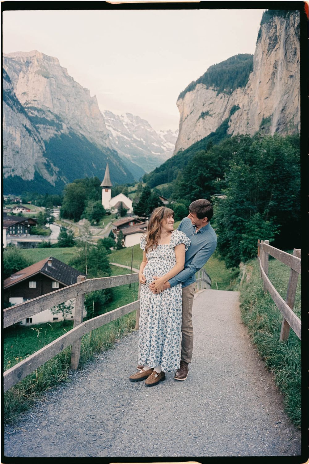 Couple embracing on a path above a Swiss alpine village with a church steeple, with steep cliffs and mountains rising behind them.