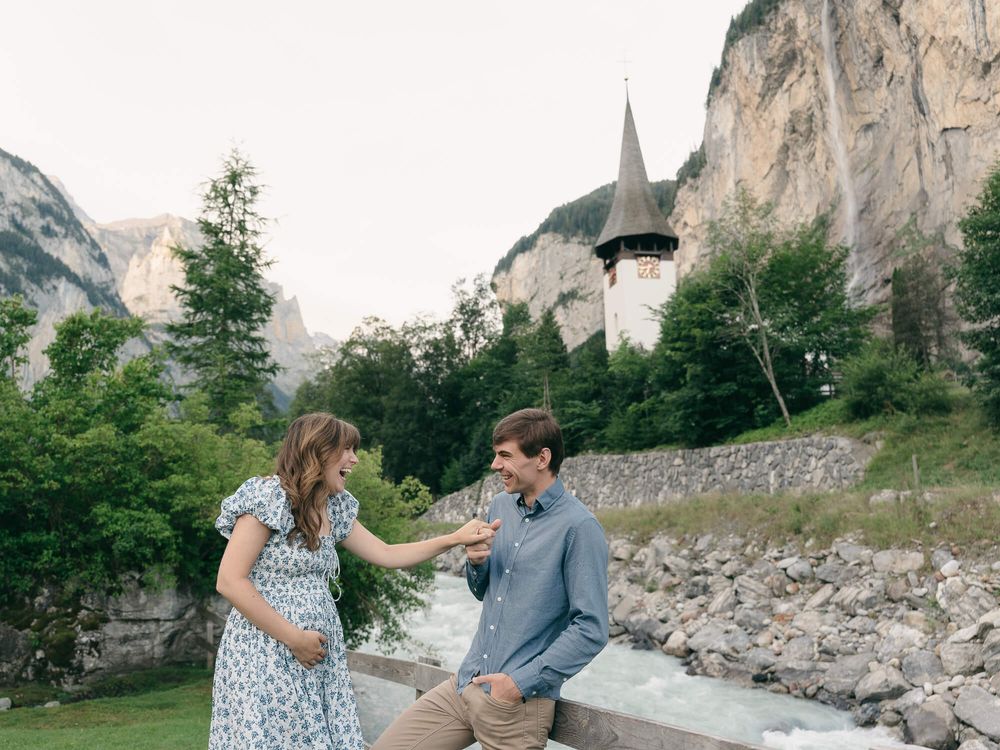 Expectant couple in a floral dress and blue shirt hold hands by a rocky stream with a stone chapel and cliffs in the Swiss Alps.