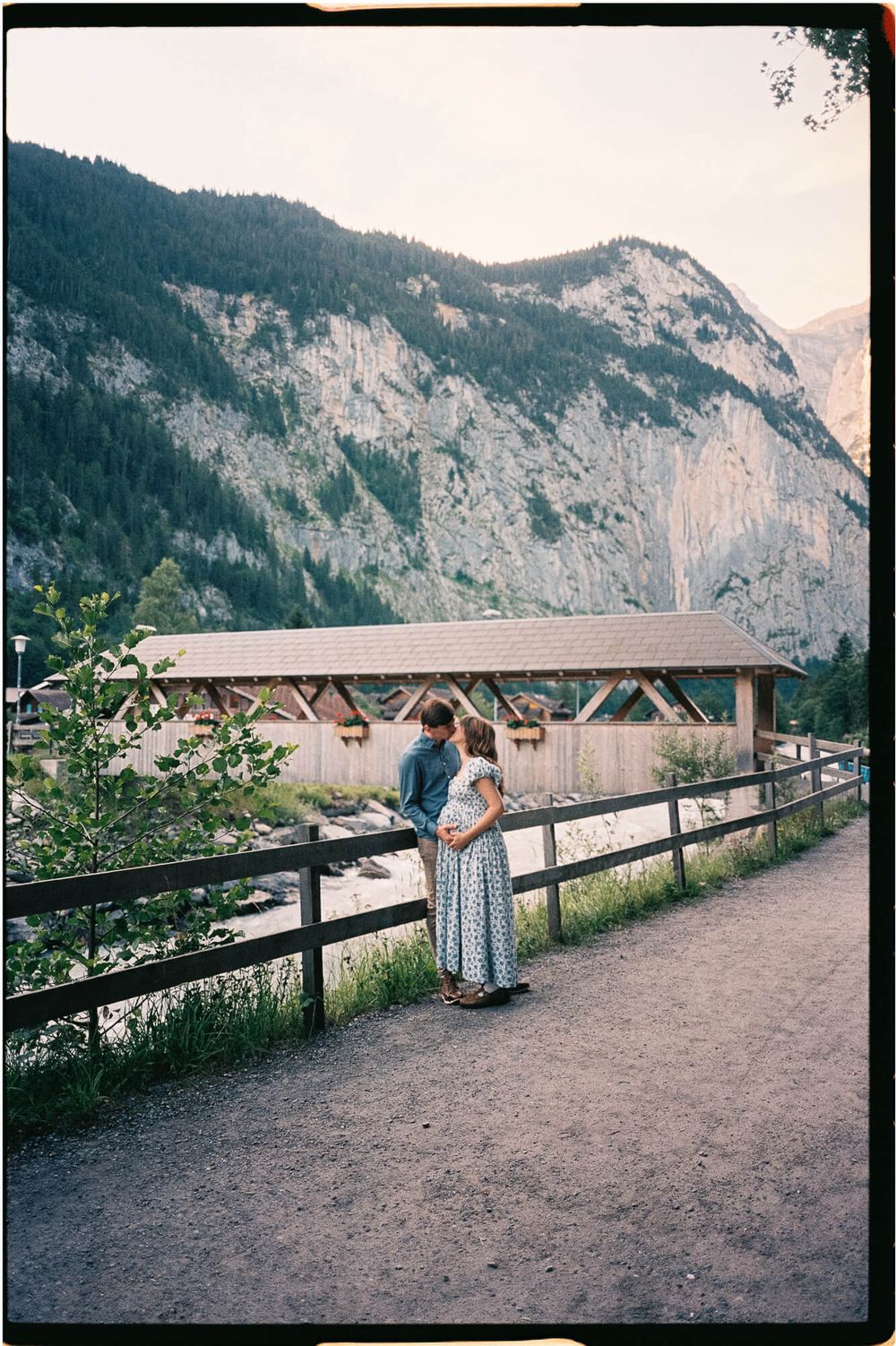 Couple embracing beside a wooden fence on a gravel path near a covered wooden bridge, with steep rocky mountains rising in the background.