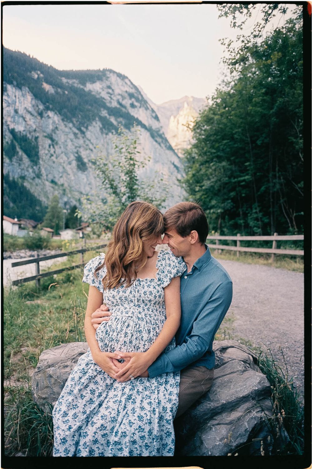 Expecting couple sits close on a boulder, the woman in a floral dress cradling her baby bump, with alpine mountains and trees behind them.