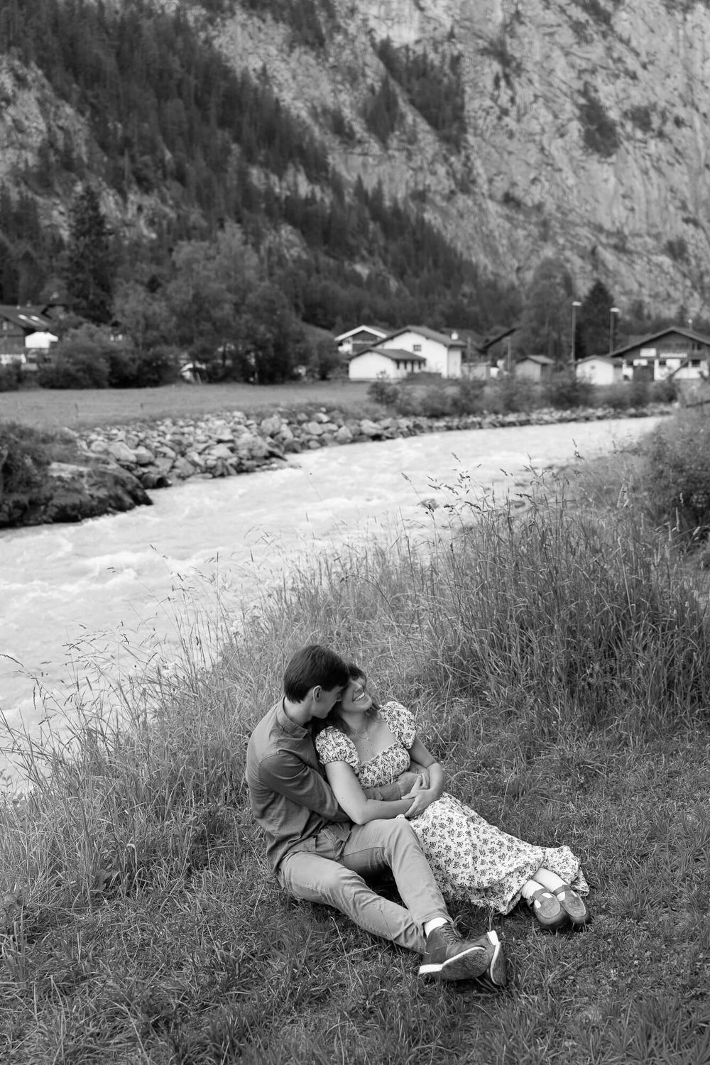 Black and white photo of a couple embracing on a grassy riverbank with a rushing stream, alpine village, and rocky mountain behind them.