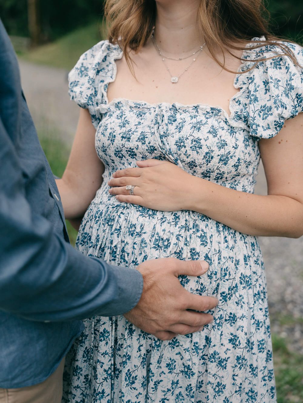 Pregnant woman in a blue floral puff-sleeve dress cradles her baby bump as a partner in a denim shirt places a hand on her belly.