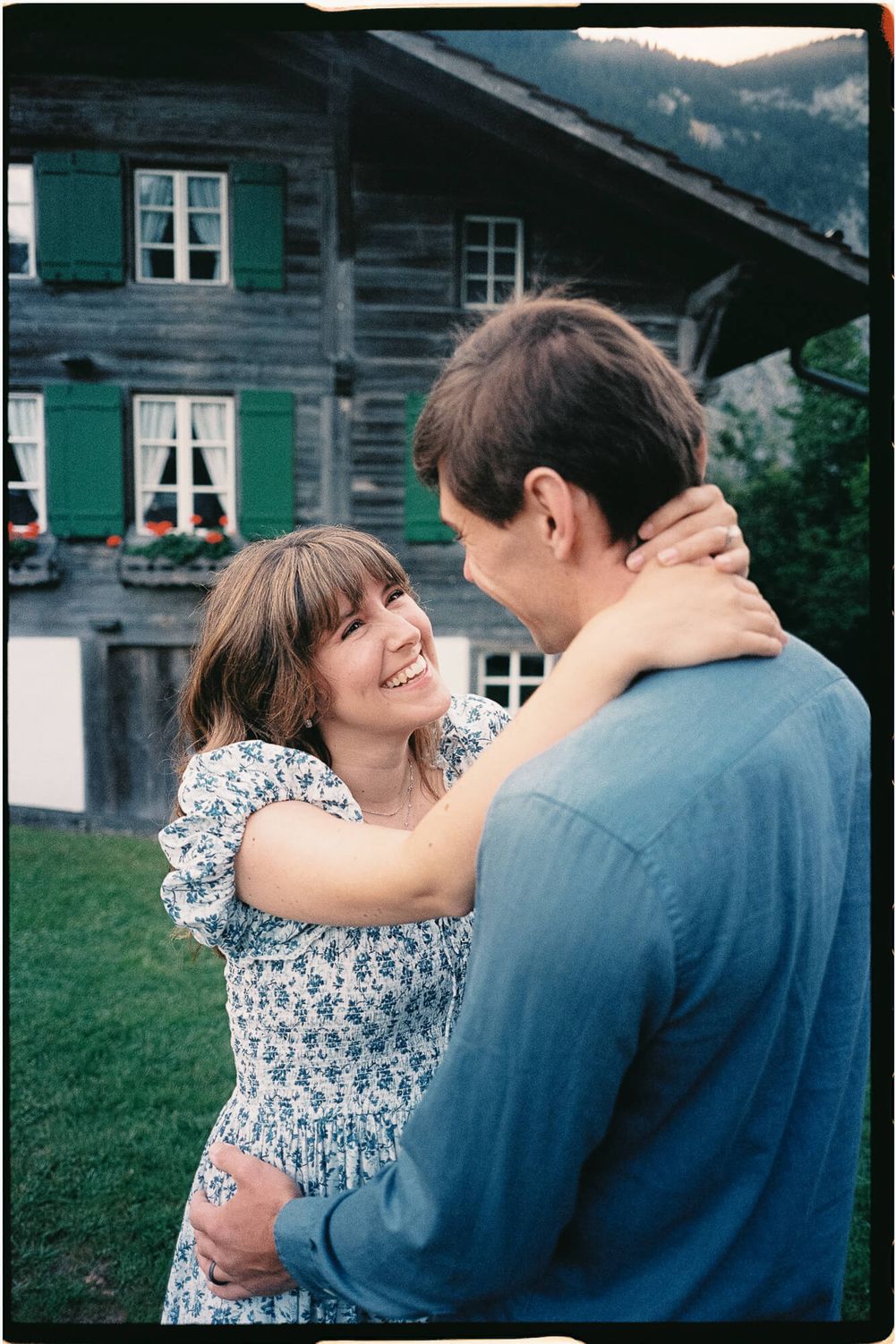 Smiling woman in a floral dress embraces a man in a blue shirt on a green lawn in front of a rustic wooden chalet with green shutters.