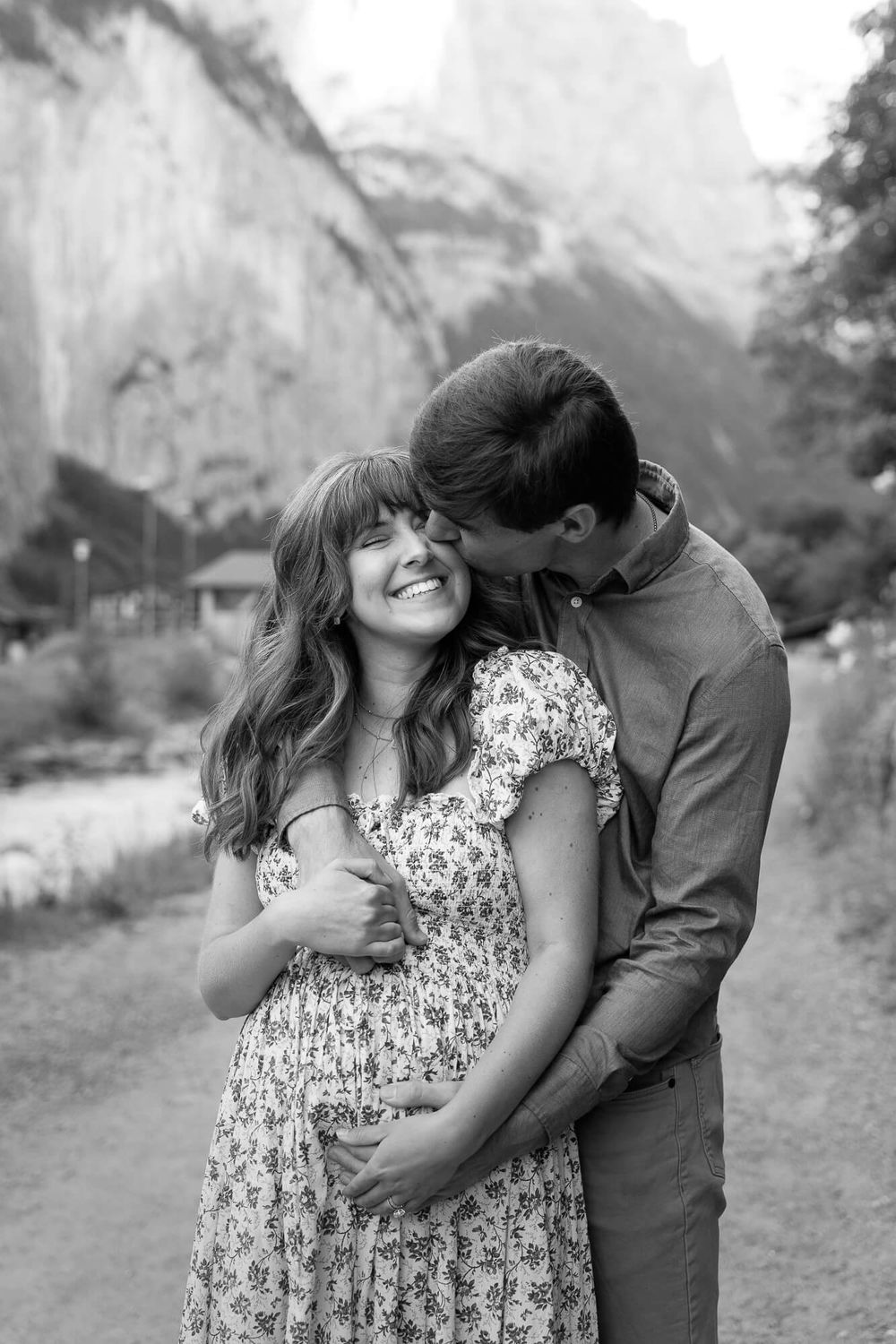 Black and white photo of a smiling pregnant woman in a floral dress embraced and kissed by her partner with cliffs in the background.