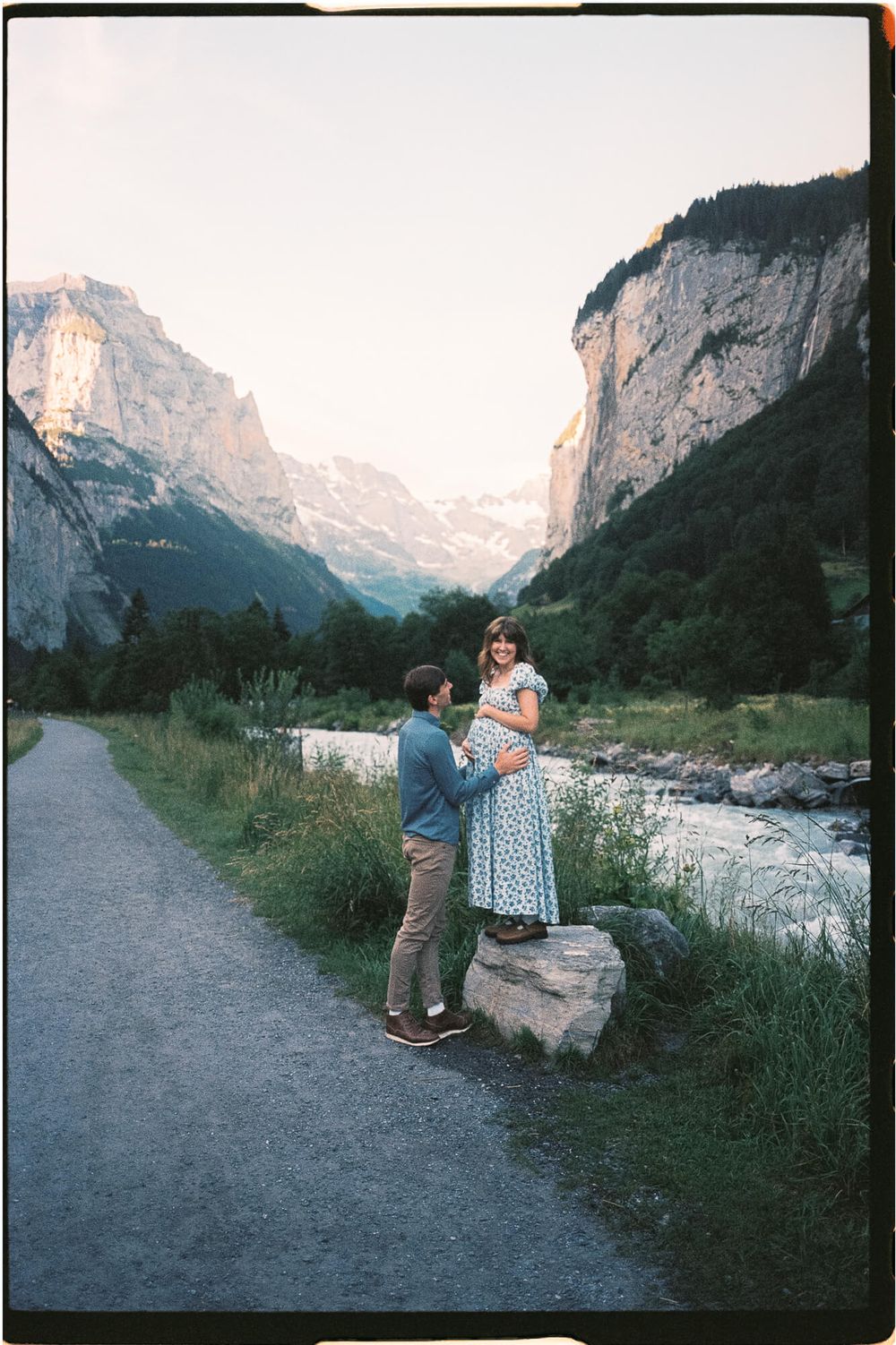 Expecting couple shares a tender maternity moment beside a glacial river with towering cliffs and alpine mountains in a scenic Swiss valley.