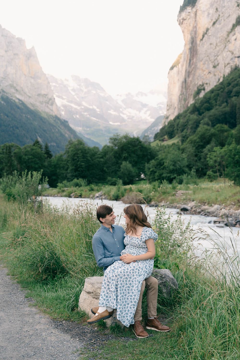 Couple sitting together on a rock beside a flowing river, surrounded by green forest and steep Alpine mountain cliffs.