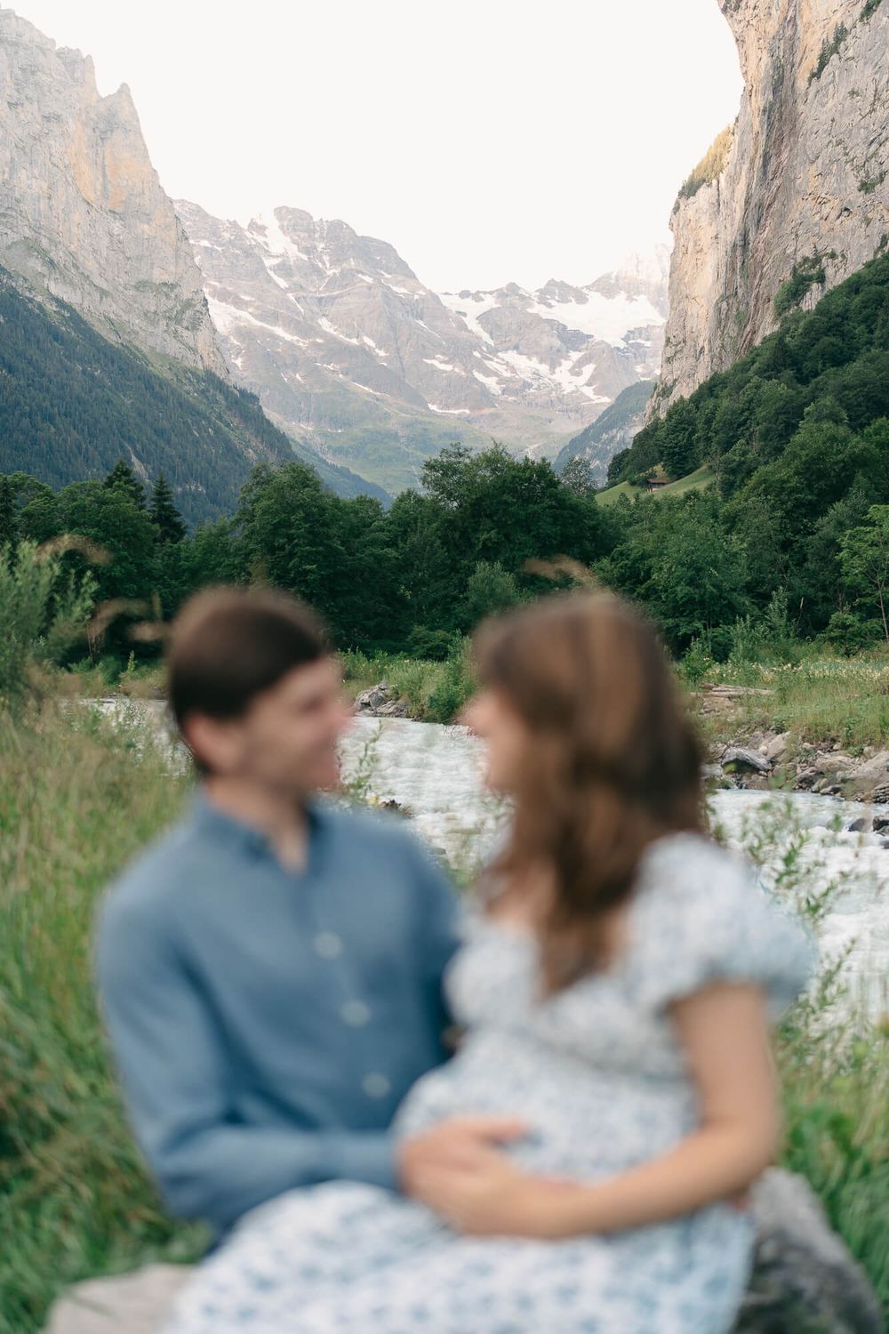 Blurred couple sits close together on a grassy bank beside a rushing stream, with towering Alpine cliffs and snowy mountains behind them.
