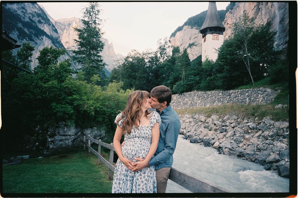 Couple embracing and kissing beside a wooden railing near a rushing mountain stream, with cliffs, green trees and a church tower behind.