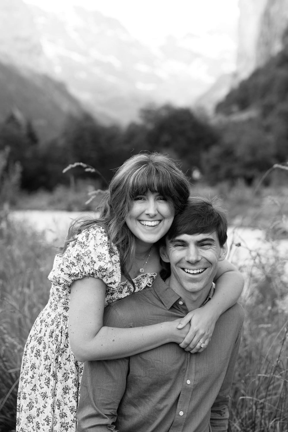 Smiling couple embracing in a grassy meadow with a river and misty mountains behind them in a black and white engagement-style portrait.