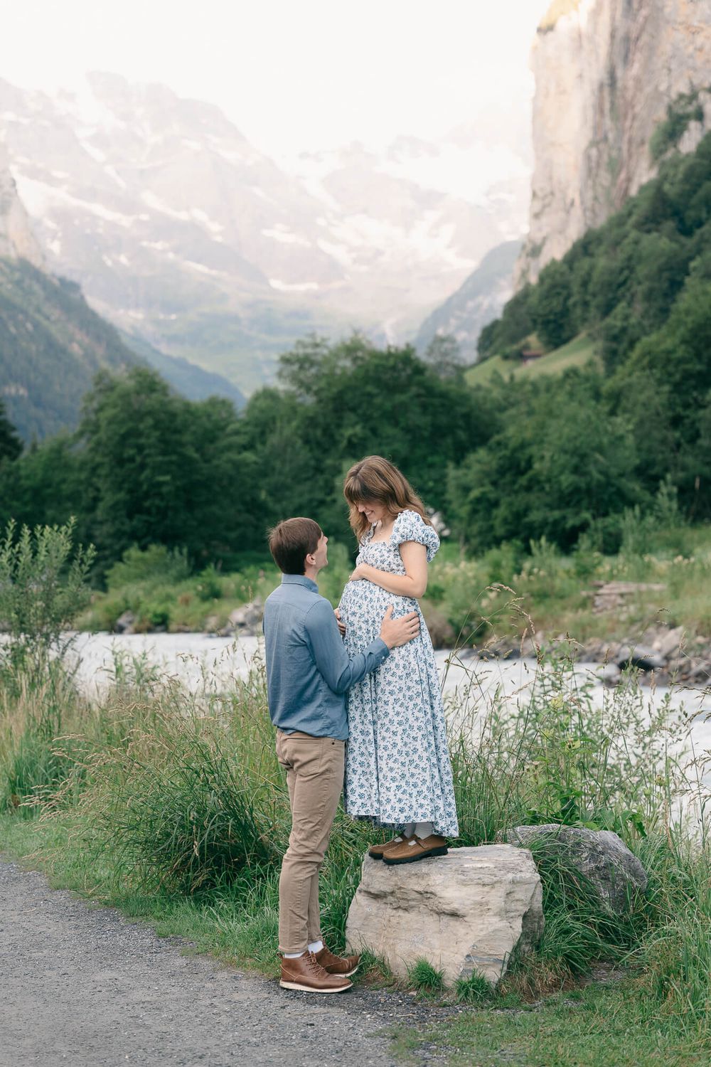 Expectant mother in a floral dress stands on a rock as her partner rests a hand on her baby bump, with Alpine mountains and a river behind.