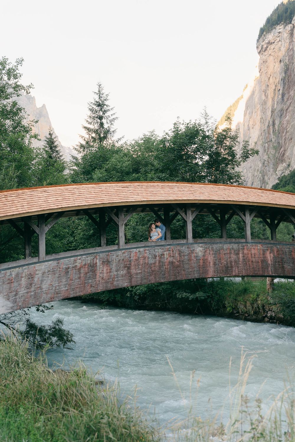 Couple embracing on a covered stone and wood bridge over a rushing alpine river, with evergreen trees and steep cliffs behind.