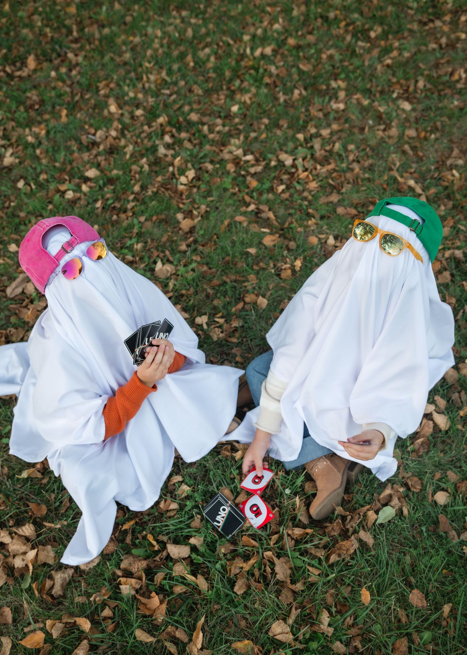 Two people in white ghost sheet costumes with pink and green beanies and sunglasses play Uno on a grassy lawn covered in autumn leaves.