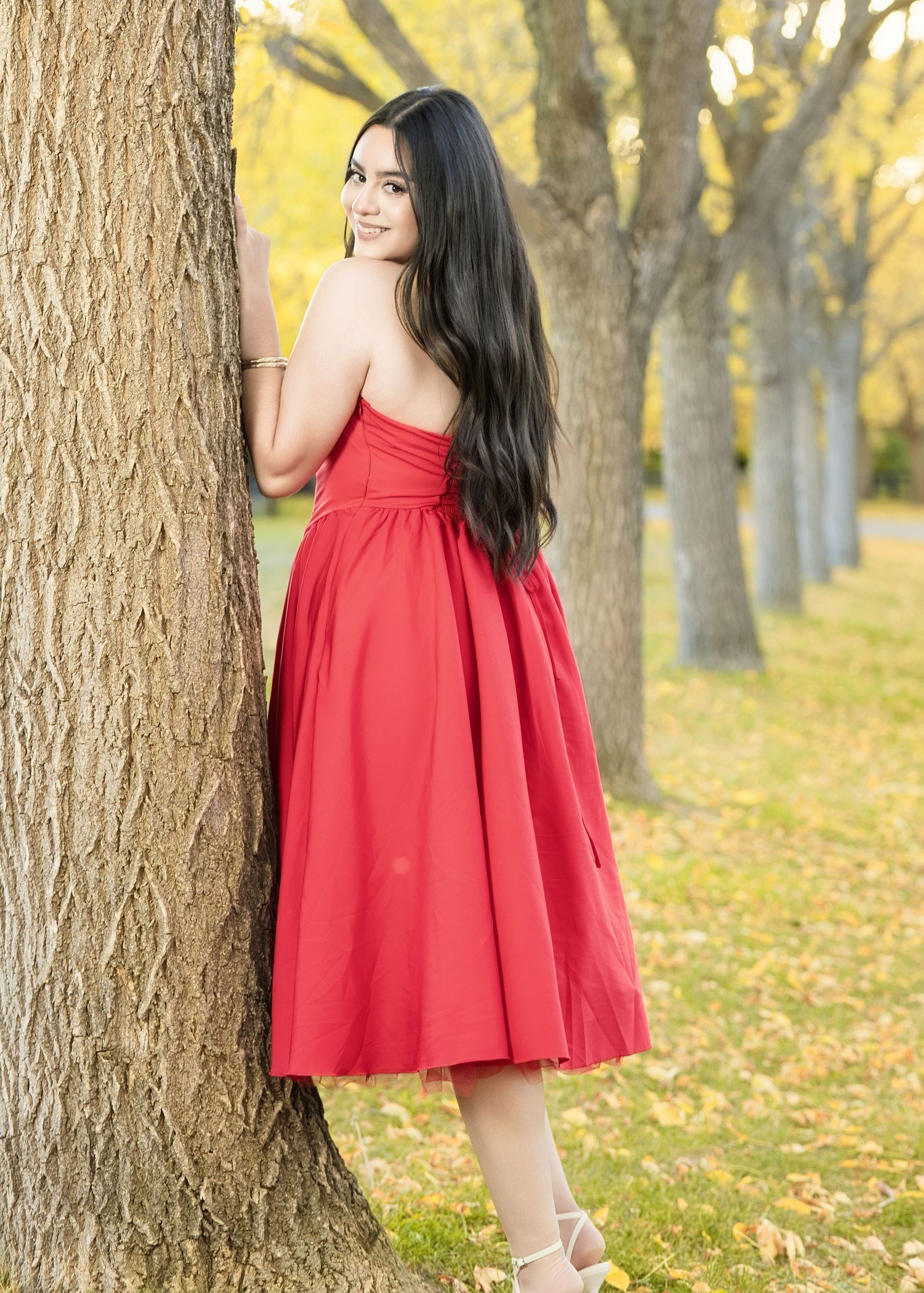 Young woman in red strapless midi dress poses by tree in autumn park with yellow foliage.