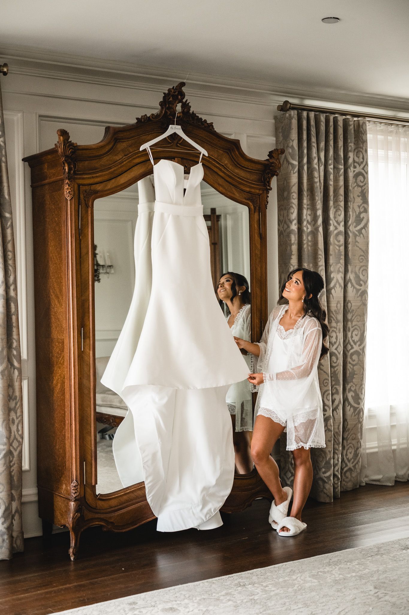 Bride in white lace robe admiring her wedding gown hanging on an ornate wooden armoire in an elegant room.