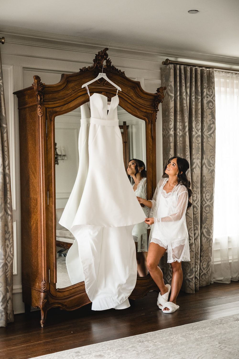 Bride in white lace robe admiring her wedding gown hanging on an ornate wooden armoire in an elegant room.
