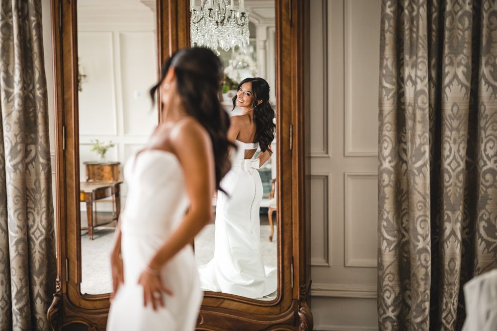Bride in white gown admires her reflection in an ornate gold mirror in an elegant room.