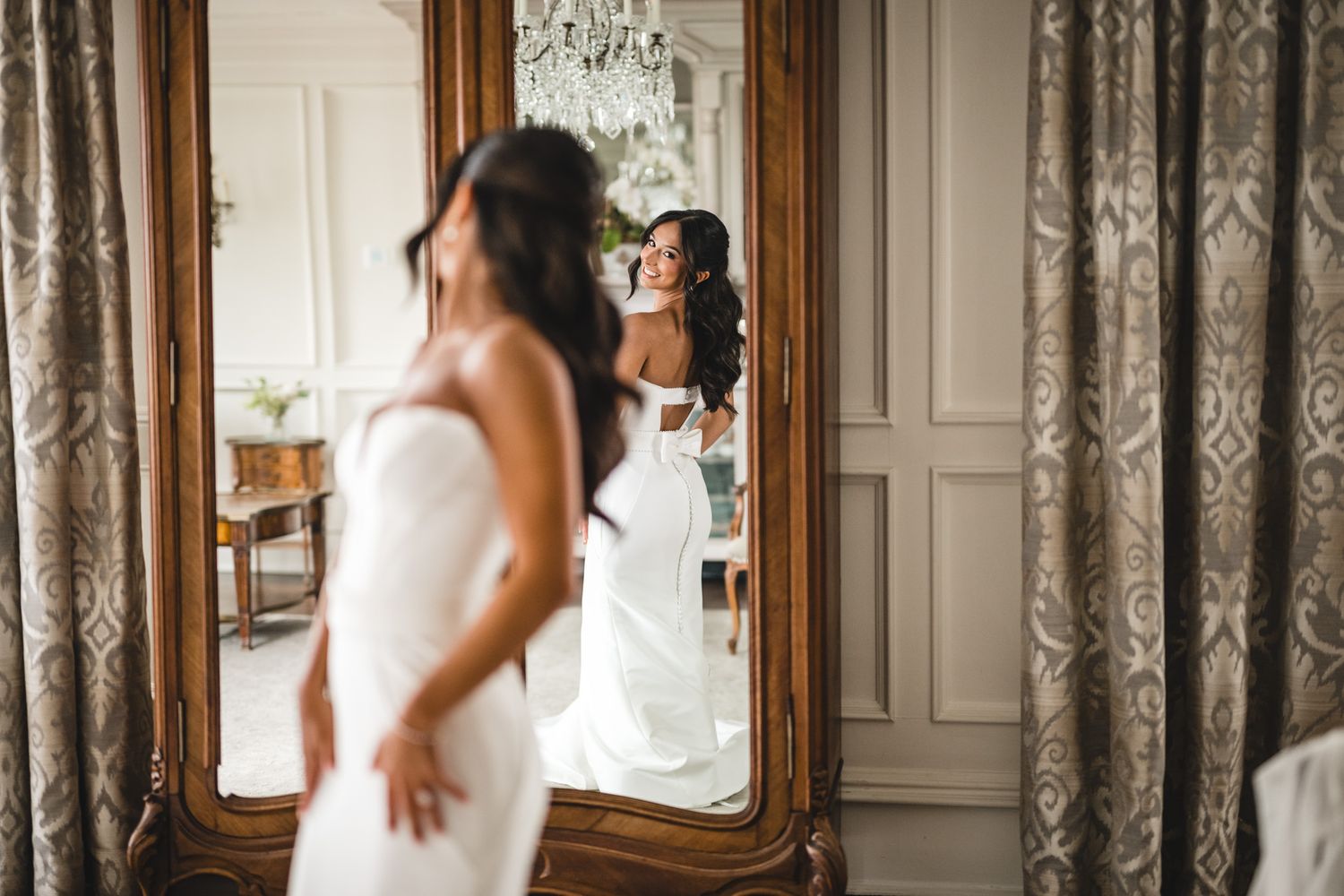 Bride in white gown admires her reflection in an ornate gold mirror in an elegant room.