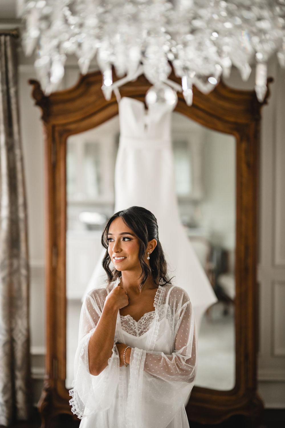Smiling bride in white lace robe stands before ornate mirror with wedding dress hanging, chandelier above.