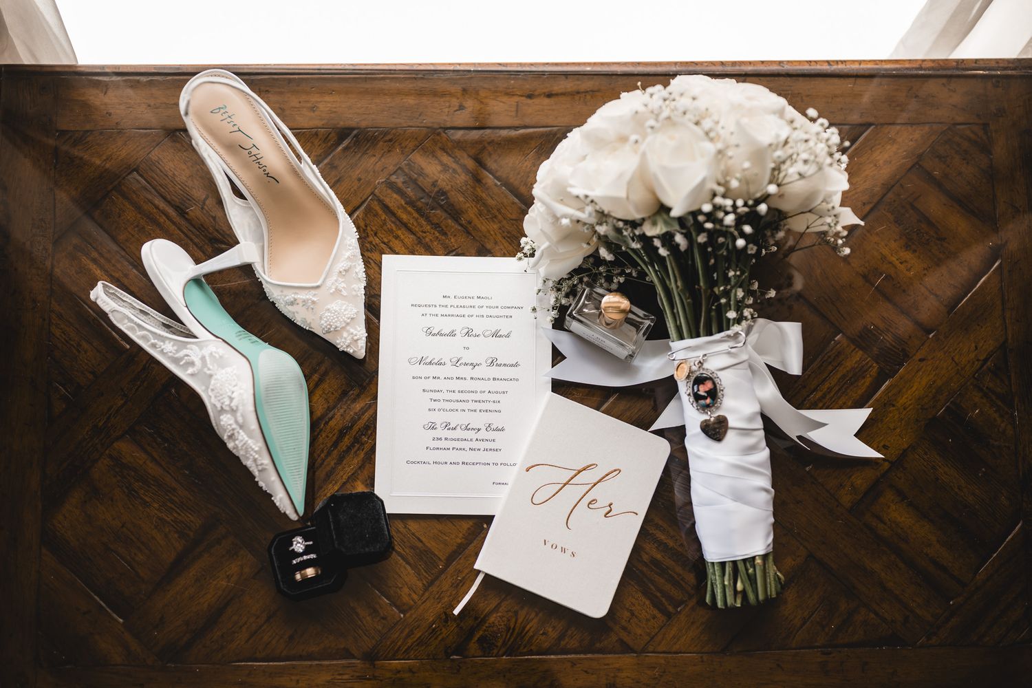 Wedding flat lay featuring white heels, floral bouquet, invitation suite, and bridal accessories on a wooden surface.