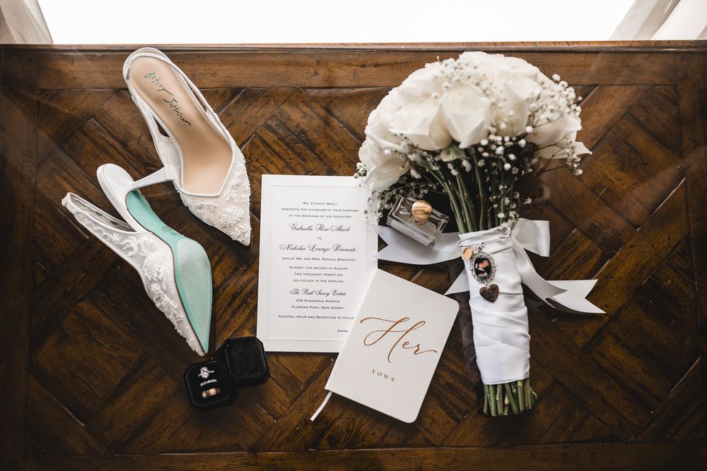 Wedding flat lay featuring white heels, floral bouquet, invitation suite, and bridal accessories on a wooden surface.