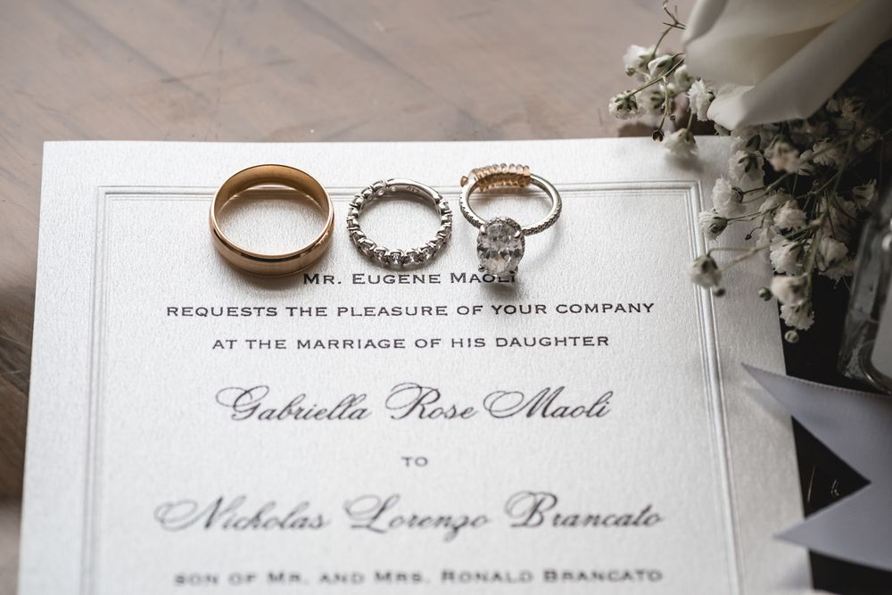 Wedding rings and bands rest on an elegant marriage invitation for Gabriella Rose Maoli and Nicholas Lorenzo Brancato.