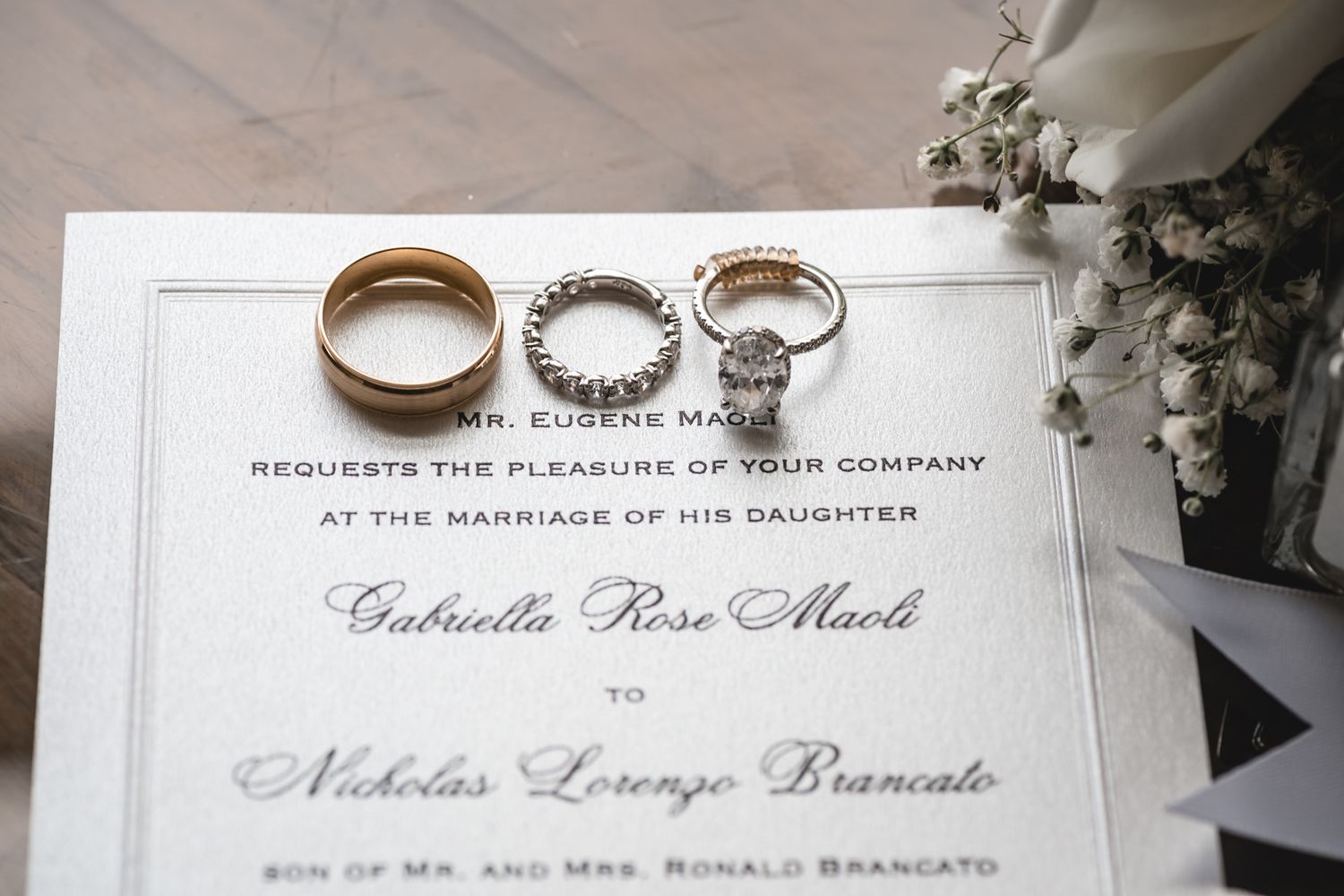 Wedding rings and bands rest on an elegant marriage invitation for Gabriella Rose Maoli and Nicholas Lorenzo Brancato.