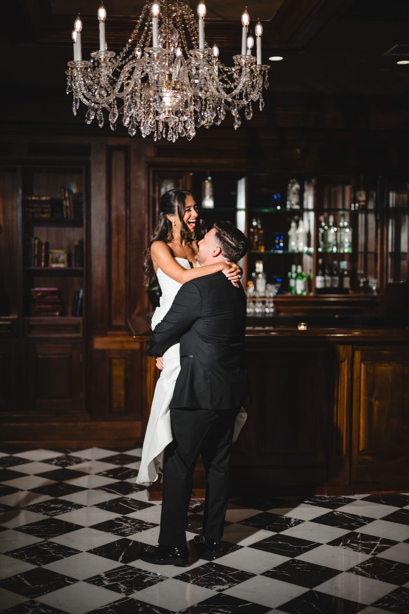 Bride and groom share a joyful embrace on a checkered dance floor beneath a crystal chandelier in an elegant venue.