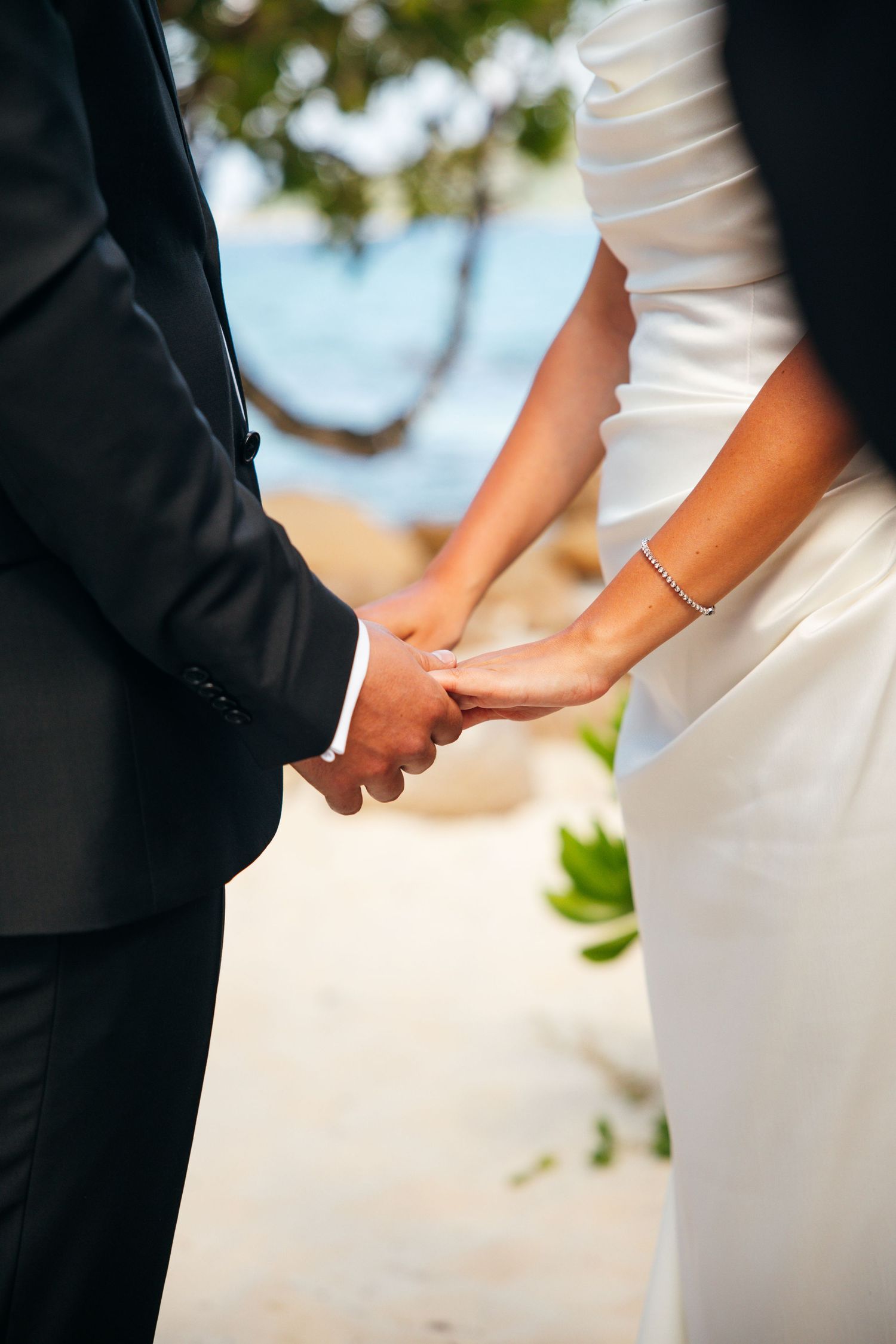 Bride and groom hold hands at tropical beach wedding ceremony in formal attire.