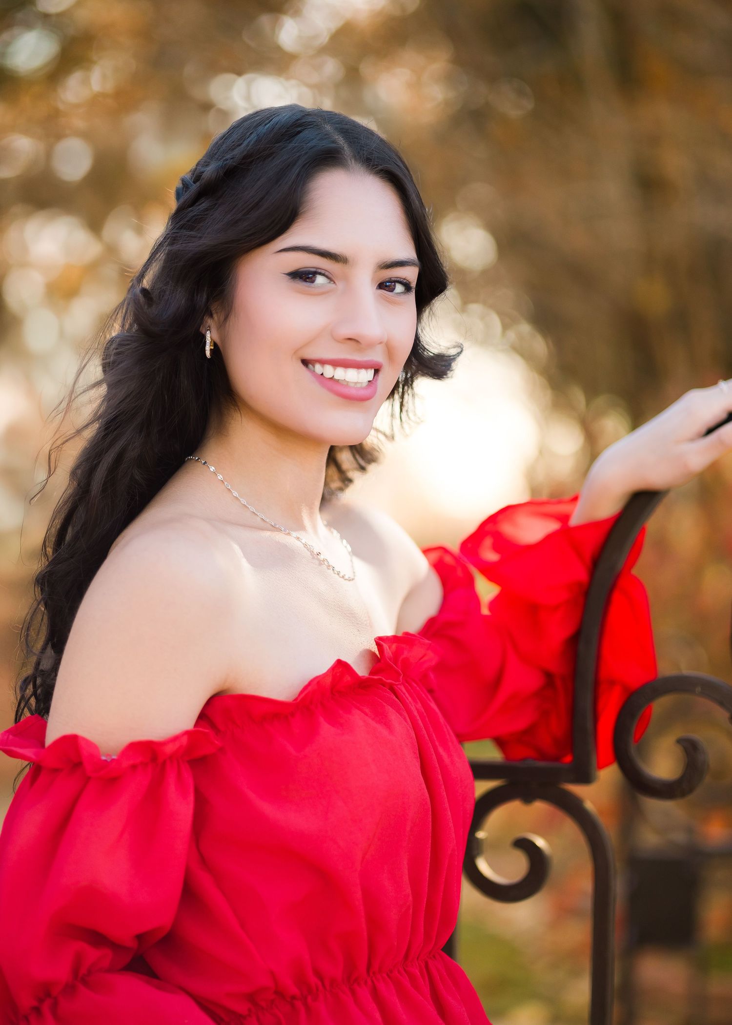 Young woman in red off-shoulder dress smiling outdoors with autumn bokeh background.